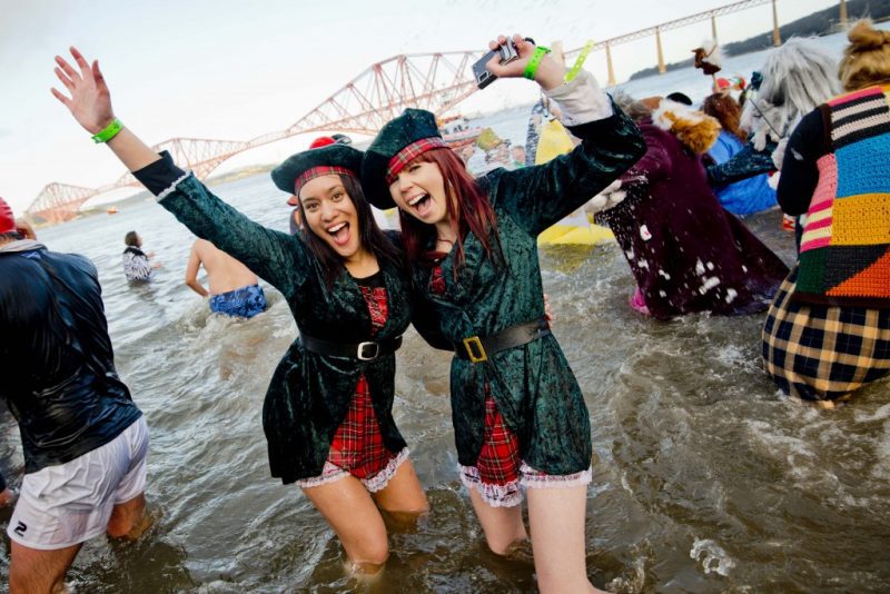 two women dressed in tartan dresses with green velvet coats and traditional scottish green hats standing in the river forth on new years day with a bridge behind them and a crowd of people swimming in fancy dress all around