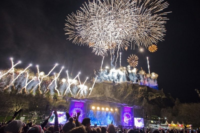 crowd in front of an outdoor stage beneath a rocky cliff with edinburgh castle on it an several white fireworks exploding in the sky above for NYE