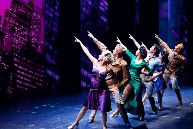 a row of men and women on a stage all pointing upwards with purple lighting, the women are wearing dresses and the men are dressed as WWII soldiers 