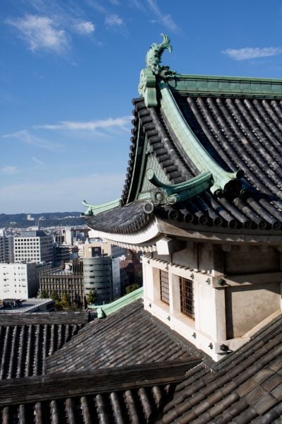Alt text: A traditional Japanese castle roof in Wakayama City with ornate architectural details, including a decorative roof ornament resembling a fish or dragon. The roof is made of dark, curved tiles and features green accents. In the background, a modern cityscape with various buildings and structures is visible under a clear blue sky.