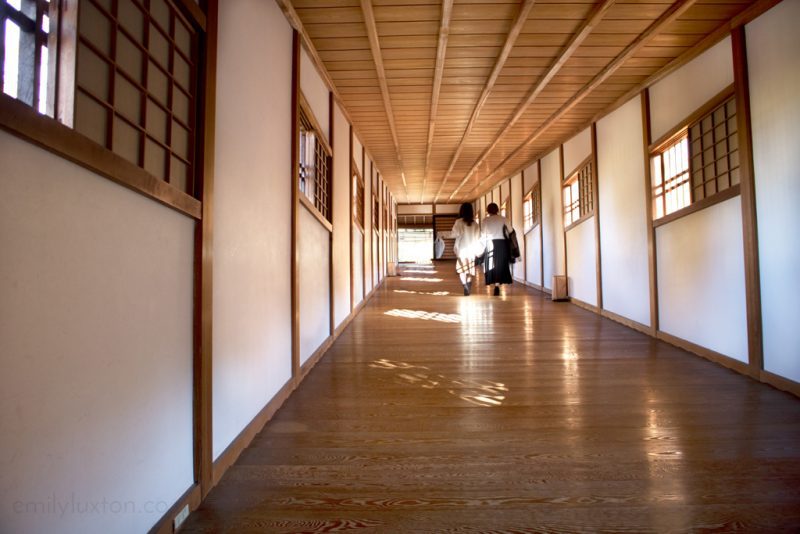 A long, wooden hallway featuring traditional Japanese architectural elements, such as wooden floors and white walls with wooden frames. The serene scene has two people walking away from the camera, with patterns of light on the floor from the windows, reflecting a calm, orderly ambiance.