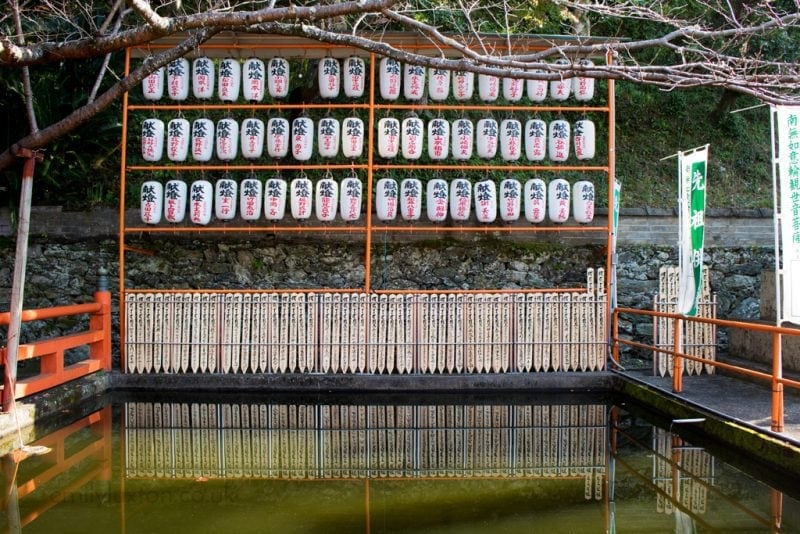Traditional Japanese lanterns and wooden plaques displayed at a shrine or temple. The lanterns, arranged in three rows, feature Japanese characters. Below them, wooden plaques with inscriptions are hung, set against a stone wall. A body of water in the foreground reflects the lanterns and plaques, with a red railing and tree branches framing the scene.