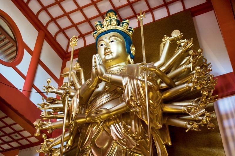 A large, golden statue of a multi-armed deity inside a temple or shrine. The statue has numerous arms extending outward, each holding different objects or making various gestures. It is adorned with intricate details, including a crown and ornate clothing. The setting appears to be indoors, with a red and white ceiling structure in the background. Wakayama City Travel Guide Japan