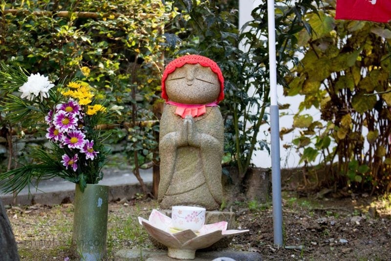 Alt text: A stone statue of a figure in a prayer position, wearing a red knitted hat and bib. In front of the statue, there's a lotus-shaped holder with a cup. To the left, a green vase holds a bouquet of white, yellow, and purple flowers. The background features green foliage and a flagpole with a red flag.