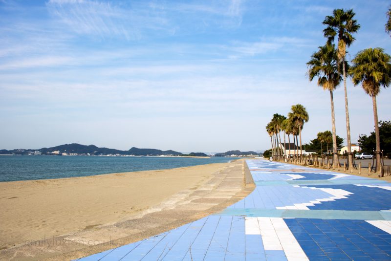 A scenic coastal area with a sandy beach on the left and a tiled promenade on the right. The promenade features a blue and white pattern and is lined with tall palm trees. In the background, there are hills and a body of water, likely an ocean or sea. The sky is clear with some light clouds. The beach is in Wakayama City Japan.