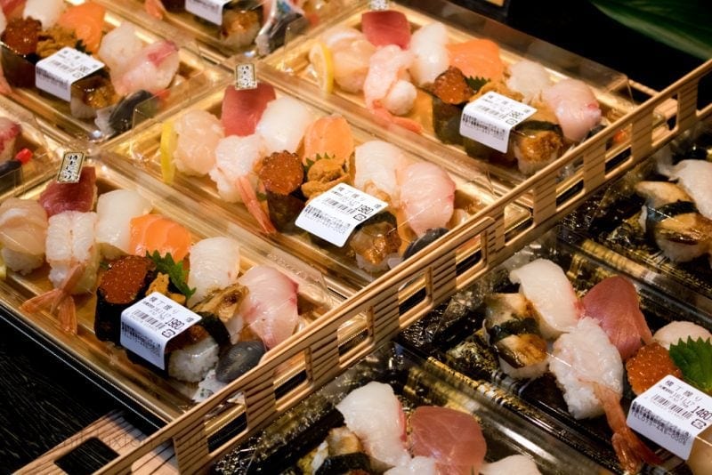 Multiple trays of assorted sushi neatly arranged in a display. Each tray contains a variety of sushi pieces, including nigiri with different types of fish and seafood like tuna, salmon, shrimp, and roe. The trays have clear lids and labels with barcodes. At the fish market in Wakayama City Japan