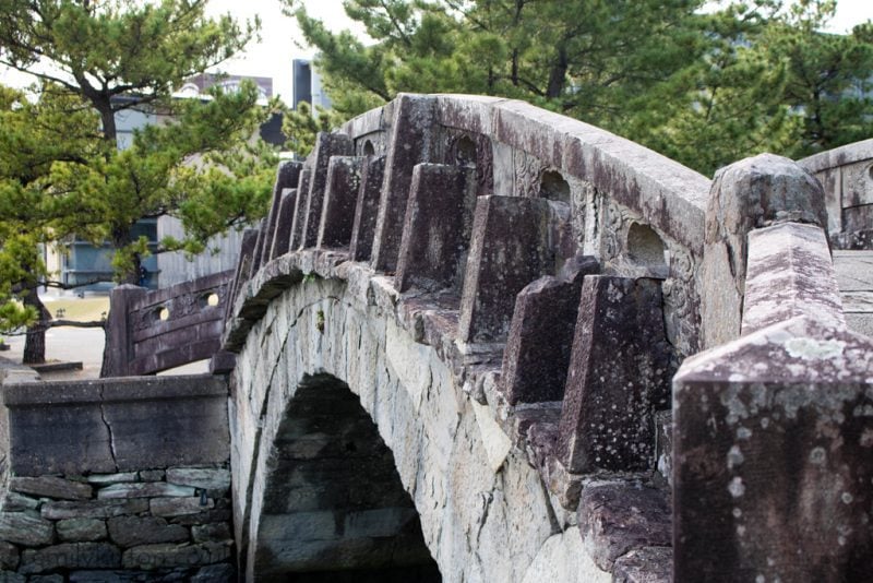 A stone bridge with an arched structure featuring vertical stone supports. The bridge shows signs of weathering and lichen growth, indicating its age. It's set in a natural environment with trees in the background. The design reflects traditional architectural techniques. Wakayama City Japan travel guide.