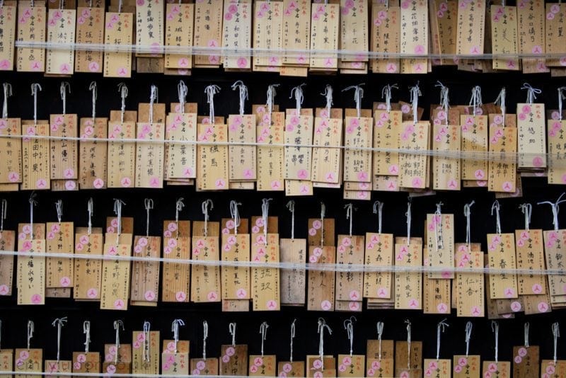 Wooden plaques, known as "ema," hanging in a Shinto shrine in Japan. The plaques are rectangular, with various prayers and wishes written on them in Japanese characters. Each plaque is tied with a string and hung on horizontal bars.