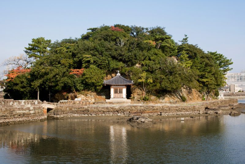A small traditional Japanese shrine structure, painted white with a tiny door and a grey tiled roof on a small island surrounded by calm water. The island, covered in dense greenery and autumn-colored trees, is connected to the mainland by a stone bridge or pathway.