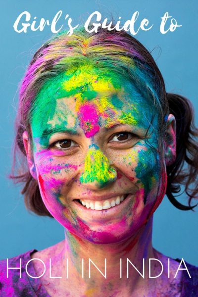 Close up of a woman's face, she has white skin and short brown hair and is smiling at the camera, and her face and hair are covered with different coloured paint. The text over the image reads: Girl's Guide to Holi in India