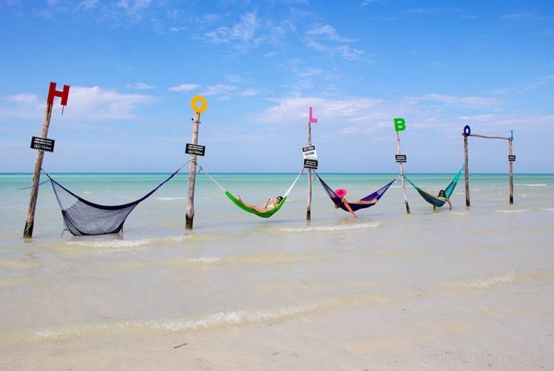 6 wooden poles in a line in the sea with different coloured hammocks strung between each pole and a letter on top of each pole spelling out the word Holbox. There is a girl wearing a pink floppy hat in the middle hammock. 