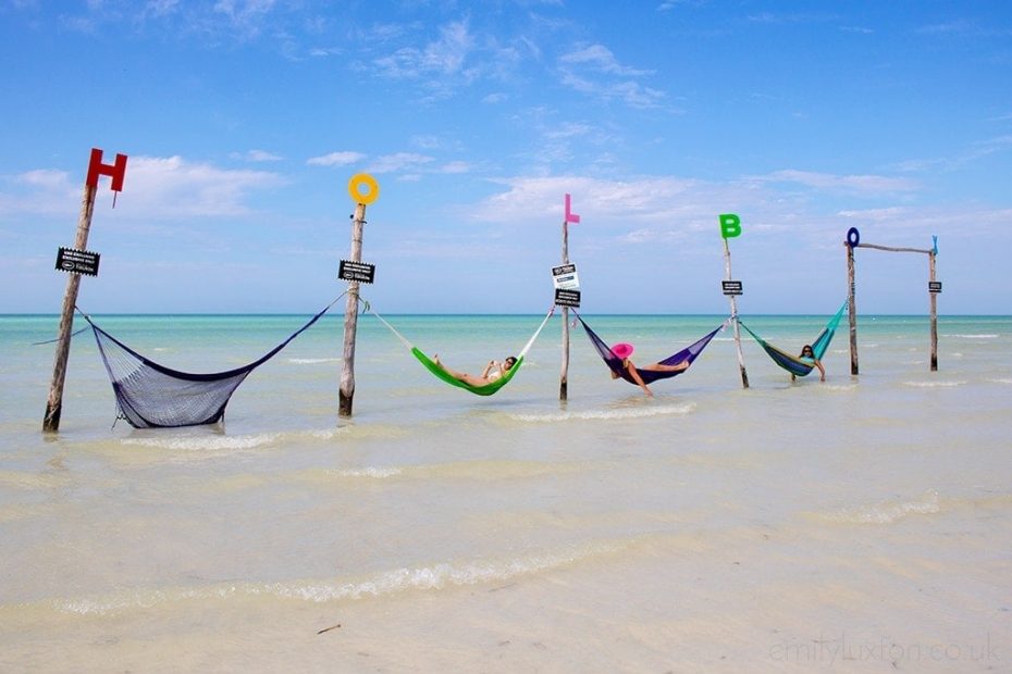 6 wooden poles in a line in the sea with different coloured hammocks strung between each pole and a letter on top of each pole spelling out the word Holbox. There is a girl wearing a pink floppy hat in the middle hammock.