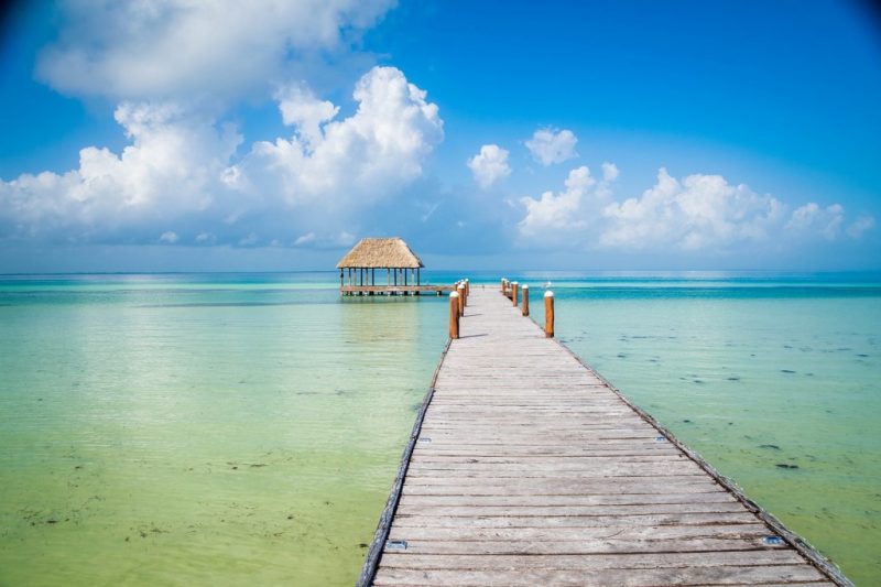 long wooden pier with a small covered section at the far end with a grass roof surrounded by shallow turquoise sea on a very sunny day with blue sky and fluffy white clouds overhead. best hotels in holbox mexico. 