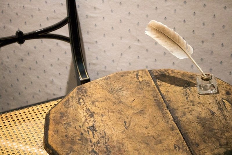 small wooden circular table with a single chair and a glass ink pot on the table with a feathered quill