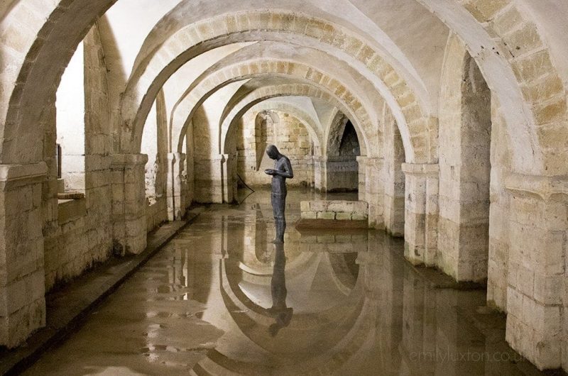 stone statue of a man looking down at his hands in a flooded crypt in winchester cathedral with the statue and stone arches overhead reflected in the water 