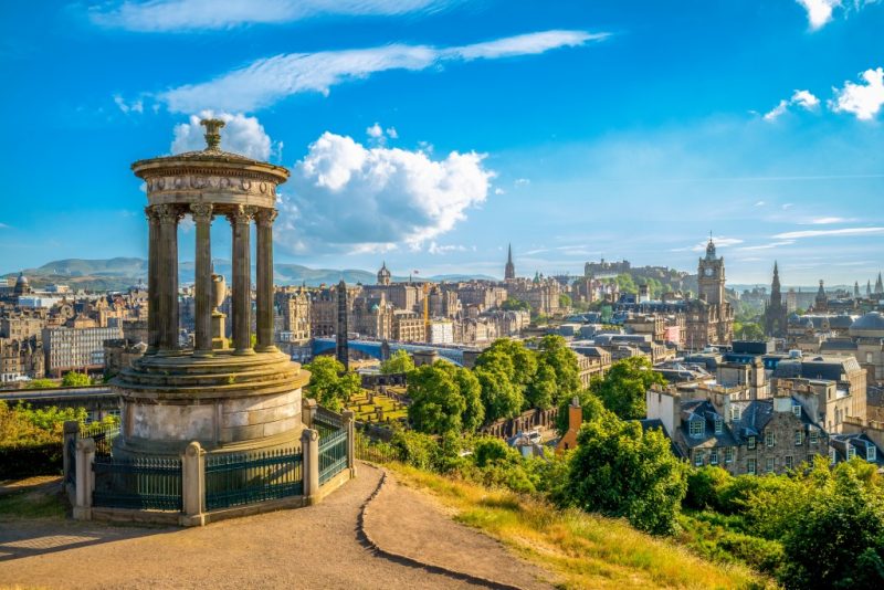 Edinburgh skyline viewed from Calton Hill on a warm sunny day with blue sky above. In the foreground is a small, circular stone monument with a domed top - the Dugald Stewart. The monument. There is a patch of greenery beyond and then the view of the city in the distance.