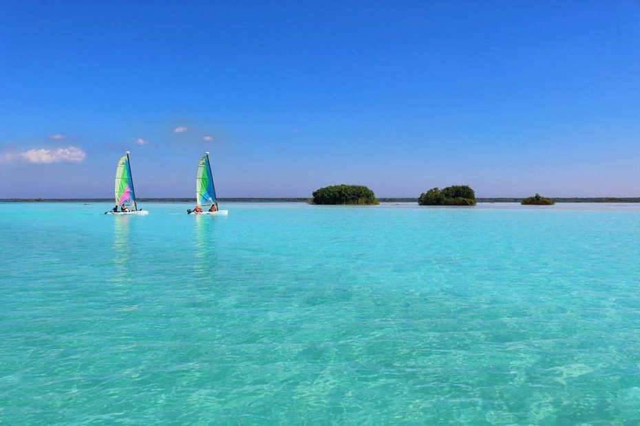 large expanse of cyan water in a lake with two tiny islands on the horizon topped with greenery and two small white sailing boats to the left in the distance with green and blue sails - how to get to Bacalar Mexico