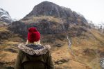 emily wearing a bright red beanie hat and a khaki green winter coat with fur trim around the hood. She is facing away from the camera and looking towards a large rocky mountain with a river running down the side in the Scottish highlands