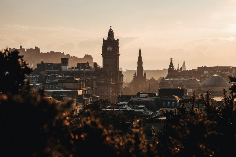 edinburgh skyline at sunset with buildings and hills silhouetted against a pale golden sky with a tall square clock tower in the centre with a pointed roof next to a church spire