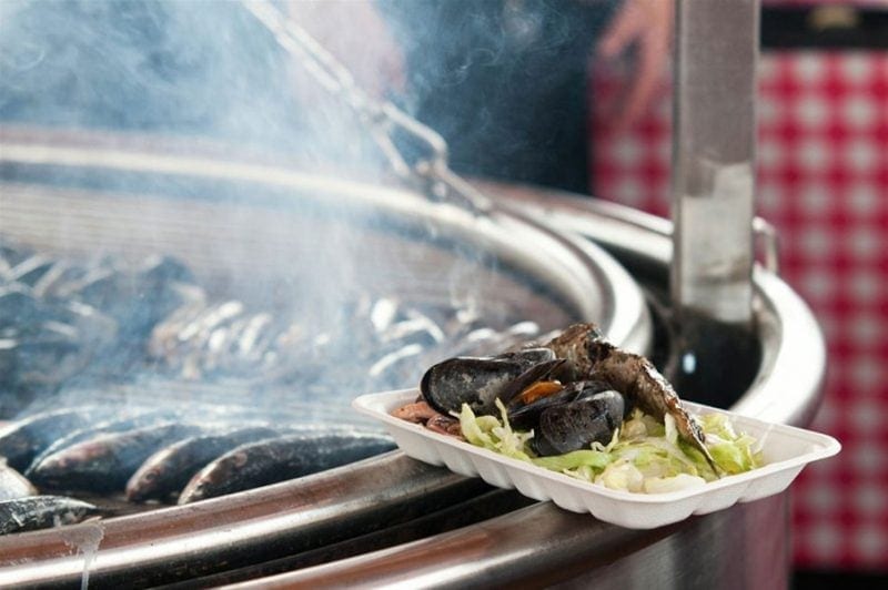 white paper tray with steamed mussels on a bed of lettuce resting on the side of a large circular pan with fish being fried on it