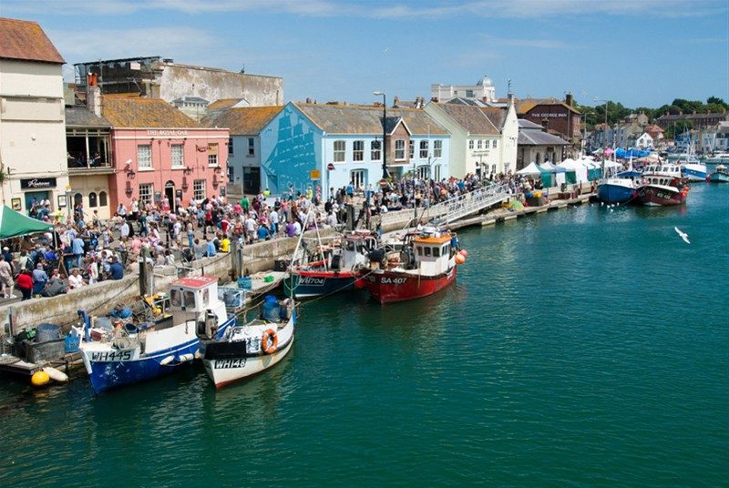 Aerial shot of Weymouth harbour on a sunny day with pastel painted buildings along the side of the harbour and a large crowd of people on the harbour wall for the Weymouth seafood festival. 