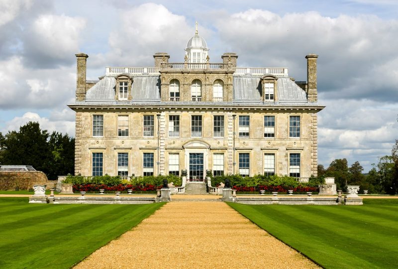 Kingston Lacy a large stately home in Dorset England built from creamy grey stone with a grey slate roof and many windows, there are neat grassy lawns in front with a gravel path between