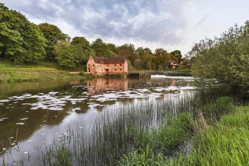 Looking across a calm, still river towards a red brick building at Sturminster Newton Mill in Dorset England