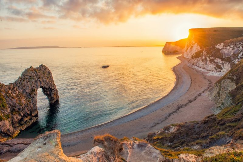 View from the edge of a grassy clifftop looking down at a bay with a curved shingle beach and a large rock archway over the sea. It is sunset and the sun is just going behind the cliffs with lens flare. 
Durdle door Dorset - UK staycation ideas