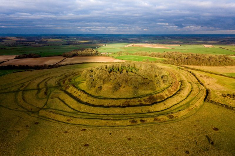 aerial view of Badbury Rings a circular iron age hill fort with a small copse of trees on the top and several grassy earth ramparts around it 