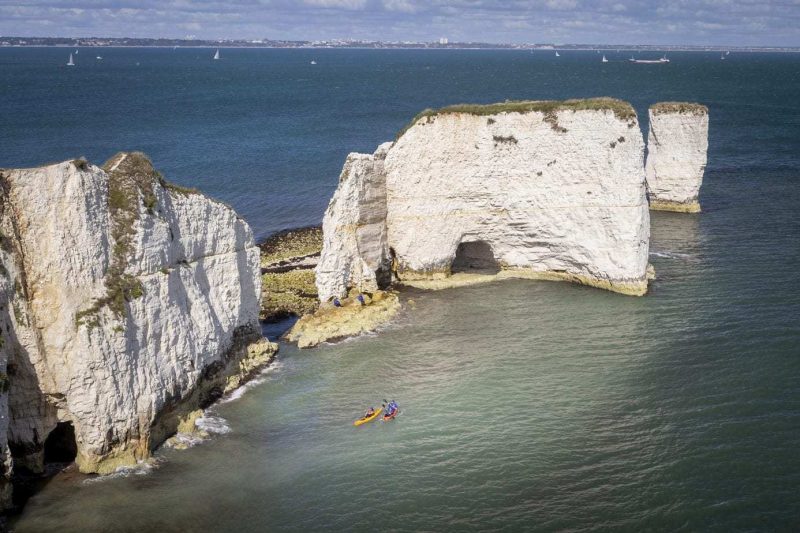 Looking down from a cliff top at several large white rock stacks in the sea with two people kayaking next to them on the jurassic coast