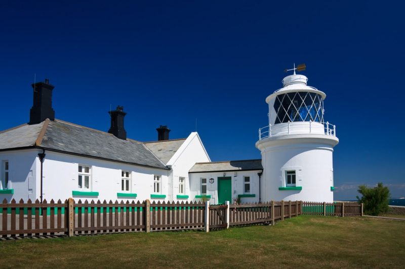 small white lighthouse building with a cottage attached, with green trim along the bottom of all the walls and windows