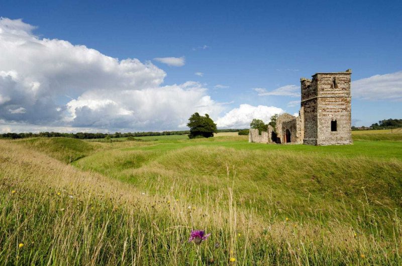small ruins of a stone church surrounded by an earth circle covered in long grass and wildflowers on a sunny summers day