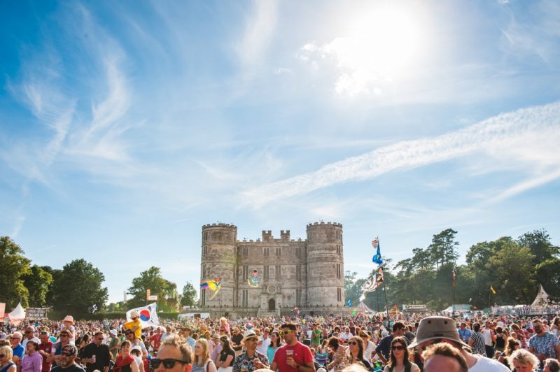 Large crowd of people at a festival holiding flags in front of a medieval grey stone castle with turretted towers on either side of the facade on a very sunny summers day