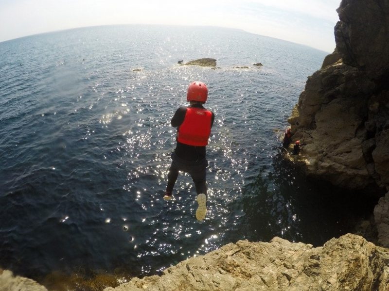 a boy in a black wetsuit with a red life jacket and red helmet jumping off a cliff into the blue sea in dorset UK