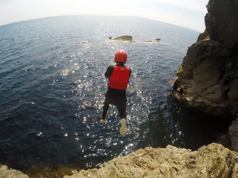 boy wearing a black wetsuite, red helmet and red lifejacket jumping away from the camera off a cliff towards the sea - unique things to do in dorset