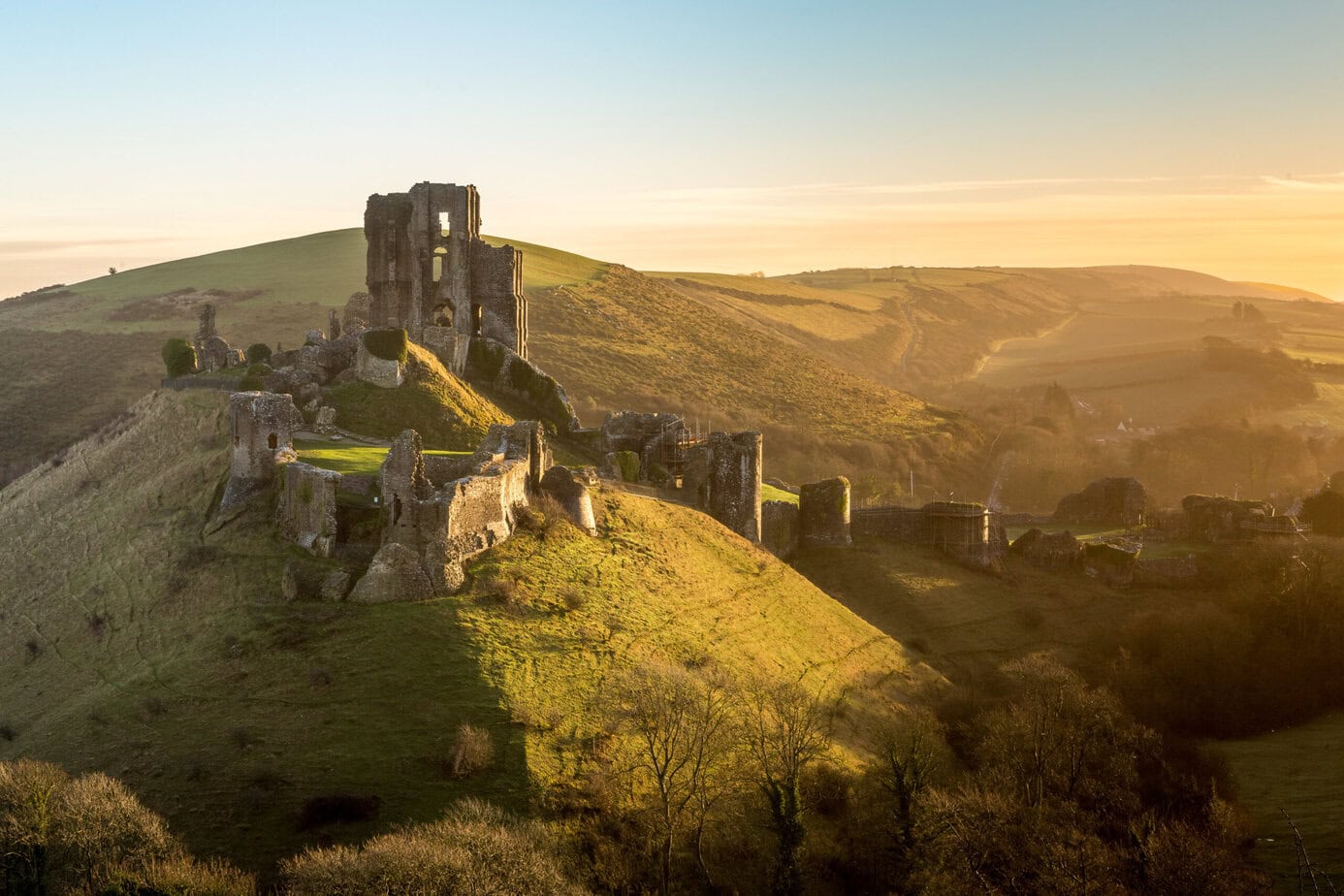 grey stone castle ruins on a hilltop surrounded by green countryside at sunrise wiht golden light