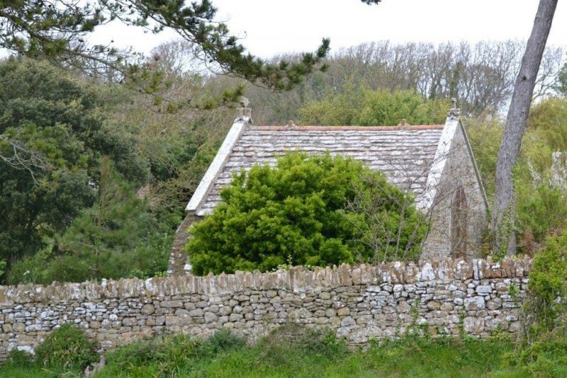 Very small single room stone church with a grey roof and grey stone walls visible behind a low stone wall and surrounded by trees