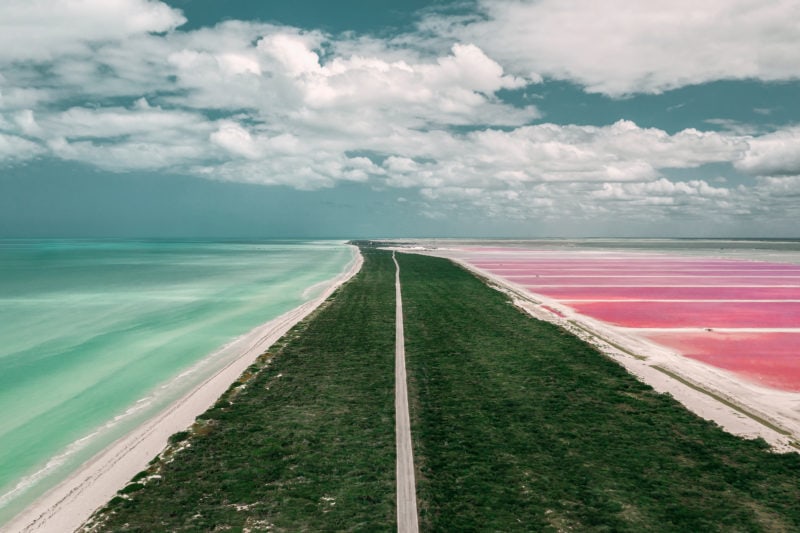 aerial view of a road running along a spit of land covered with green shrubbery, on one side is the turquoise ocean and on the right is a bright pink lake. Las coloradas in Rio Lagartos Mexico
