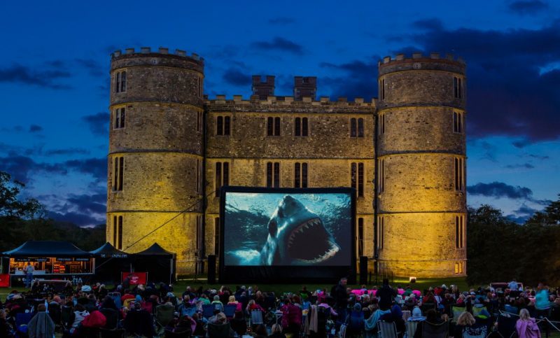 Large medieval style castle with turretted towers on either side and a big cinema screen set up on front showing a scene from JAWS with a close up of the shark 