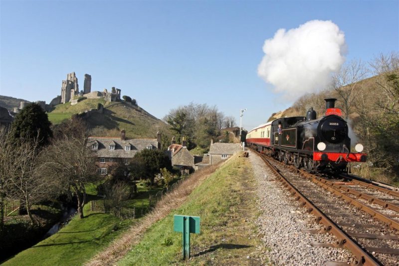 Black steam train pulling carriages running along a railway line next to a ruined castle on a hill