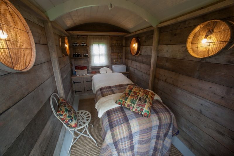 Interior of a small wooden shepherds hut with exposed wood walls and a small shelf unit at the end, in front there is a single bed with a massage fitting and a woodlen tartan blanket