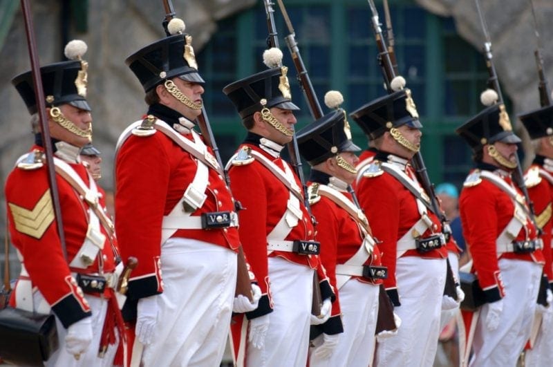 Row of men dressed in historic soldiers uniforms with red jackets, white houses and tall black hats holding rifles and facing forwards.