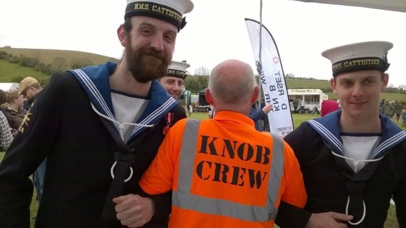 two men in navy uniform with hats that say HMS Cattistock linking arms with a man facing away from the camera in an orange high vis jacket with the words Knob Crew printed on the back. 