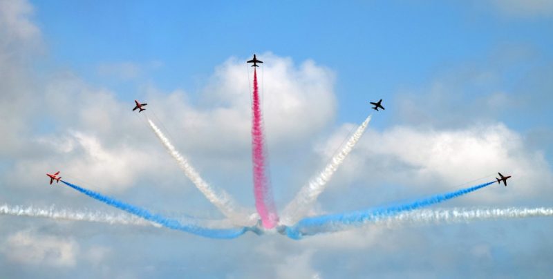 5 planes in the sky flying in different directions from the same point in a semi circular formation, the central plane has a red smoke trail, the two either side have white smoke trails and the outer two have blue smoke trails