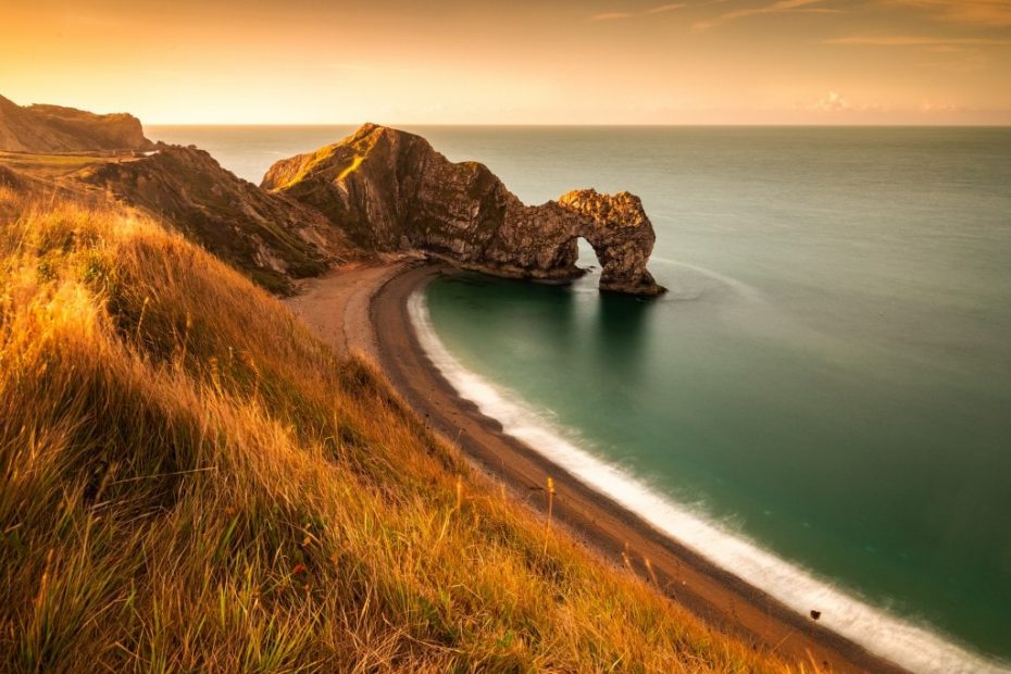 View of Durdle Door at sunset from a grassy clifftop looking down at the shingle beach and the large rock archway over the sea. Things to do in dorset