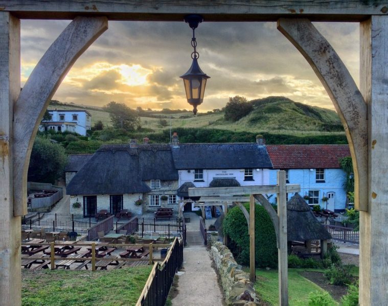 wooden gate with a lantern hanging from it with a paved path leading down towards three terraced cottages, the left hand one has grey stone walls and a thatched roof, the middle one is painted pink with a grey tiled roof and the cottage on the right is painted blue and has a red tiled roof. There are grassy hills behind and a alrge pub garden in front. Smuggler's Inn at Osmington Mills in Weymouth. 