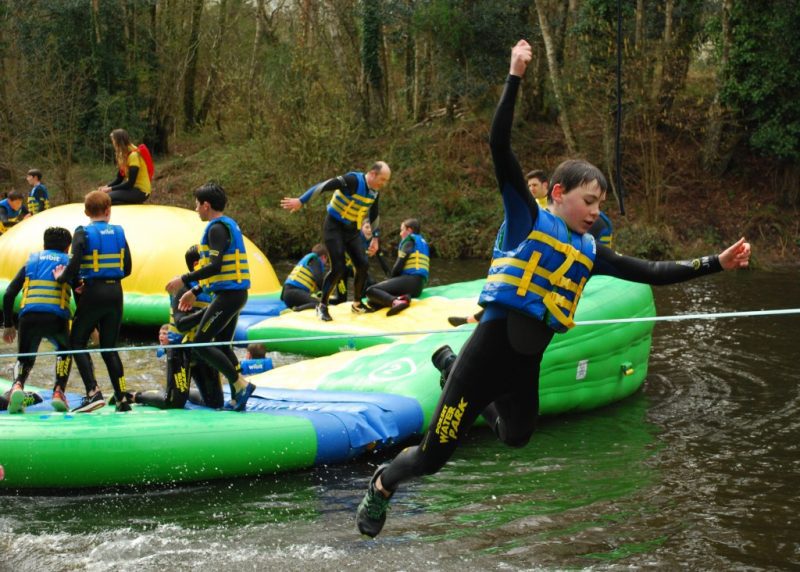 boy wearing a black wetsuit and a blue lifejacket jumping into a river in a woodland with a large green and yellow inflatable obstacle course behind him where there are many more boys wearing the same