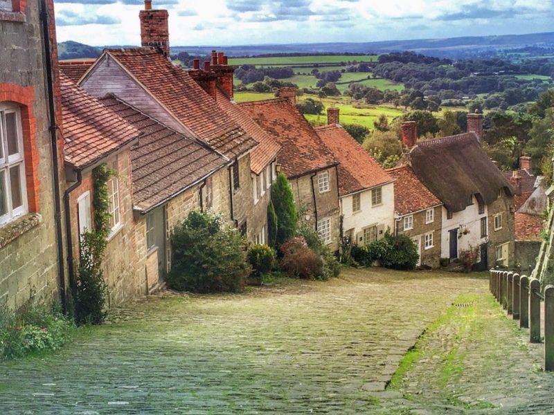 steep hill with a cobbled road and a row of terraced stone cottages with red tiled roofs with a view of green countryside and woodlands beyond