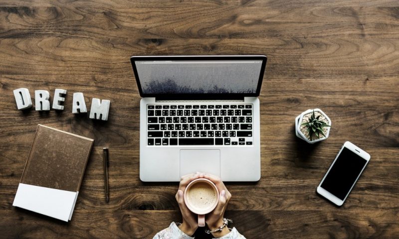 flat lay of a laptop on a large wooden table with a womans hands holding a coffee cup and a notebook, plant and phone all laid out around the laptop as well as some plastic letters spelling out the word dream. How to make money from a travel blog. 