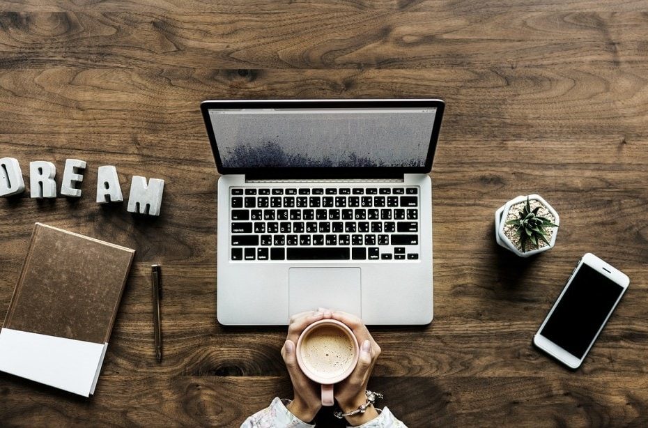 flat lay of a laptop on a large wooden table with a womans hands holding a coffee cup and a notebook, plant and phone all laid out around the laptop as well as some plastic letters spelling out the word dream. How to make money from a travel blog.